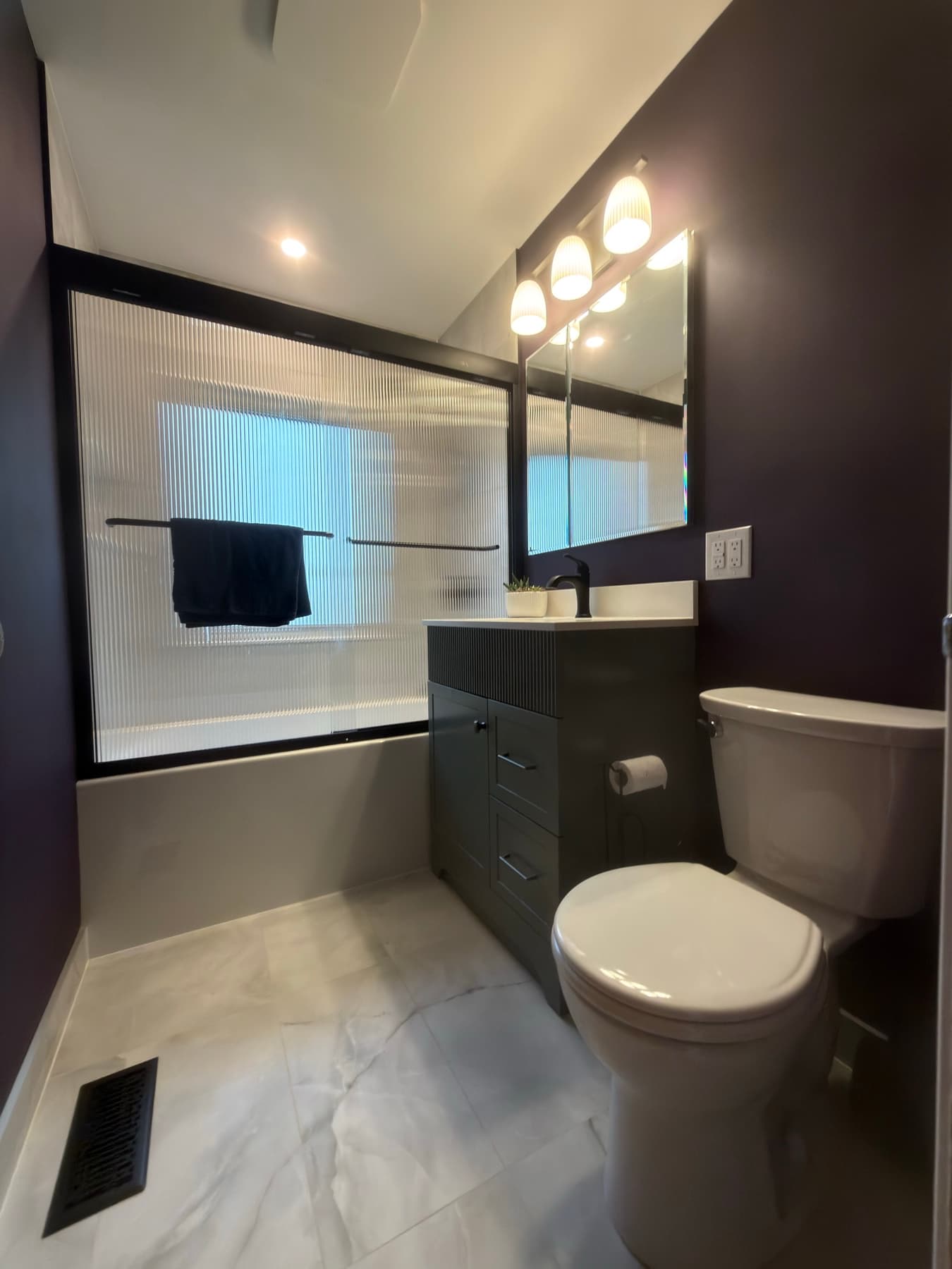 Deep plum bathroom with a dark vanity, reeded glass tub enclosure and warm vanity lighting